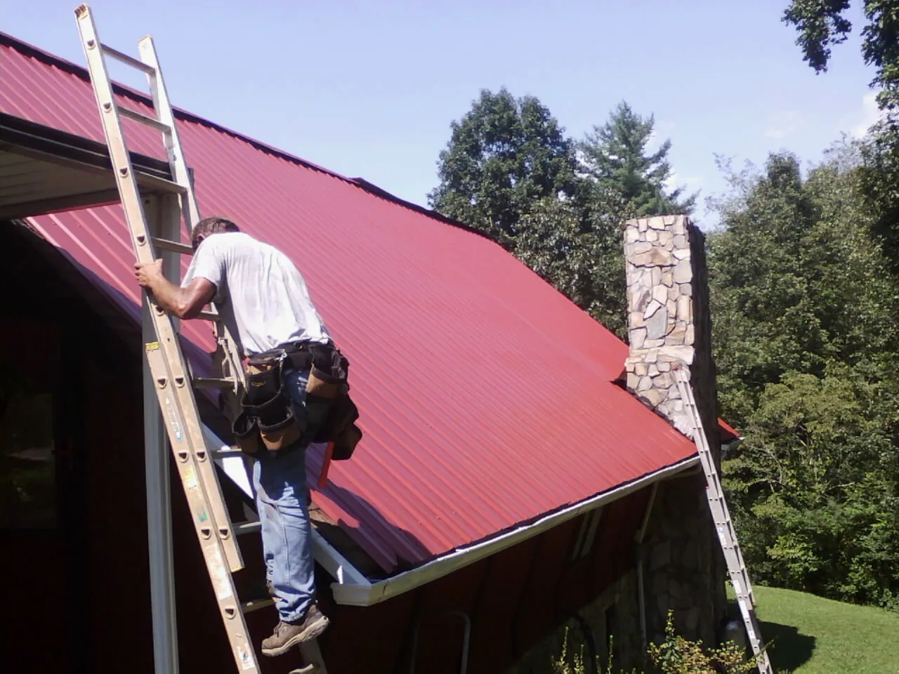 Roofer on a ladder beside a red metal roof and stone chimney — Soffit & Fascia Repair by Roofing Brothers in Danielson